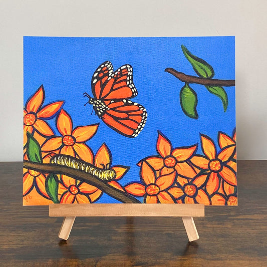 A colourful print on an easel of a painting that depicts a monarch butterfly in mid-flight against a bright blue sky. Below the butterfly, a yellow caterpillar crawls along a branch. The branch extends into vibrant orange flowers on the left and a green chrysalis hanging on the right. NZ Art Print, surreal, giclée . Flutter to Bloom