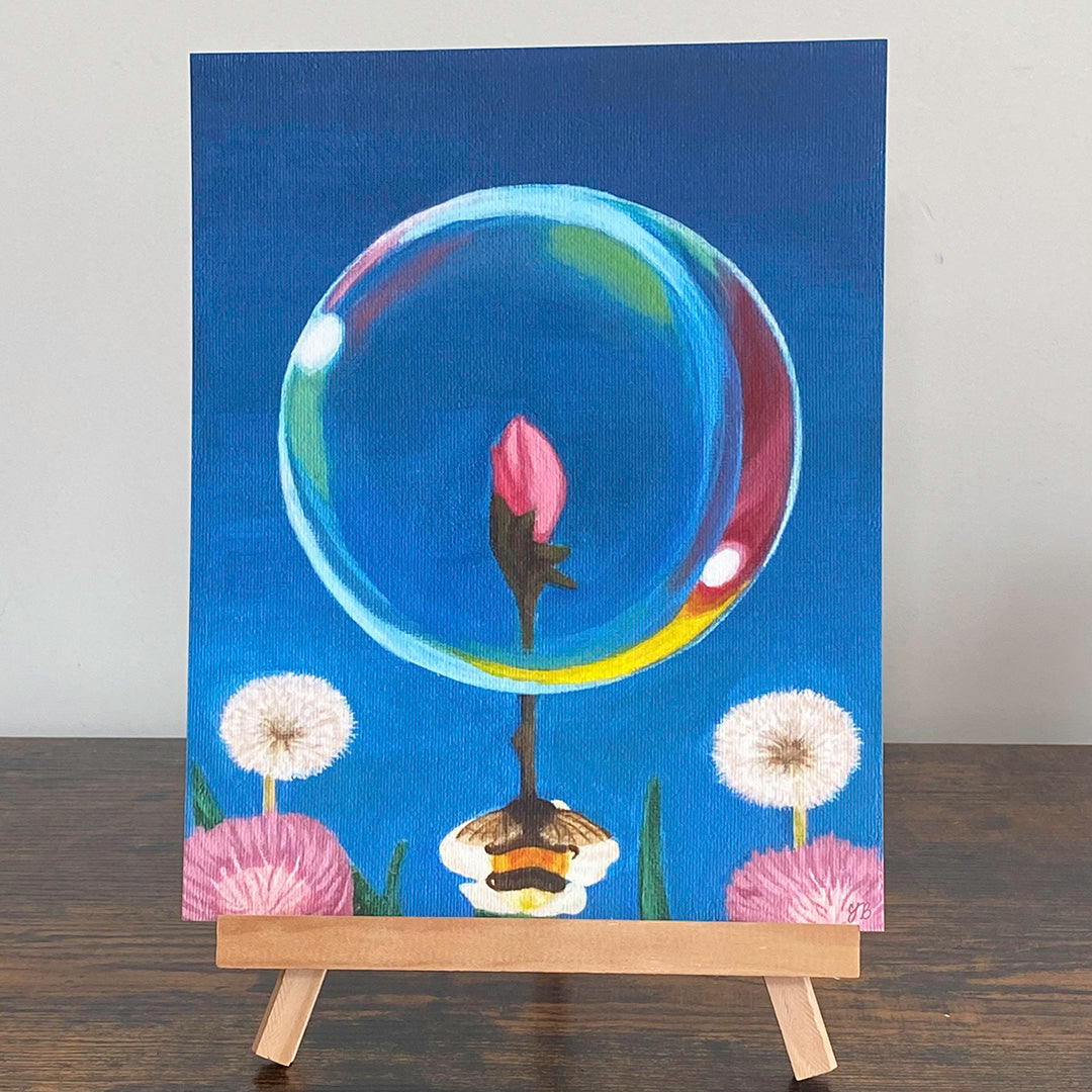 A print of a painting on an easel.  A translucent blue bubble containing a single pink flower bud. The sphere rests on top of a green stem with a bumble bee reflected below. Two white dandelions and a purple flower are visible in the background against a vibrant blue sky. Surrealism Wall Art Prints, giclée. Flutter to Bloom